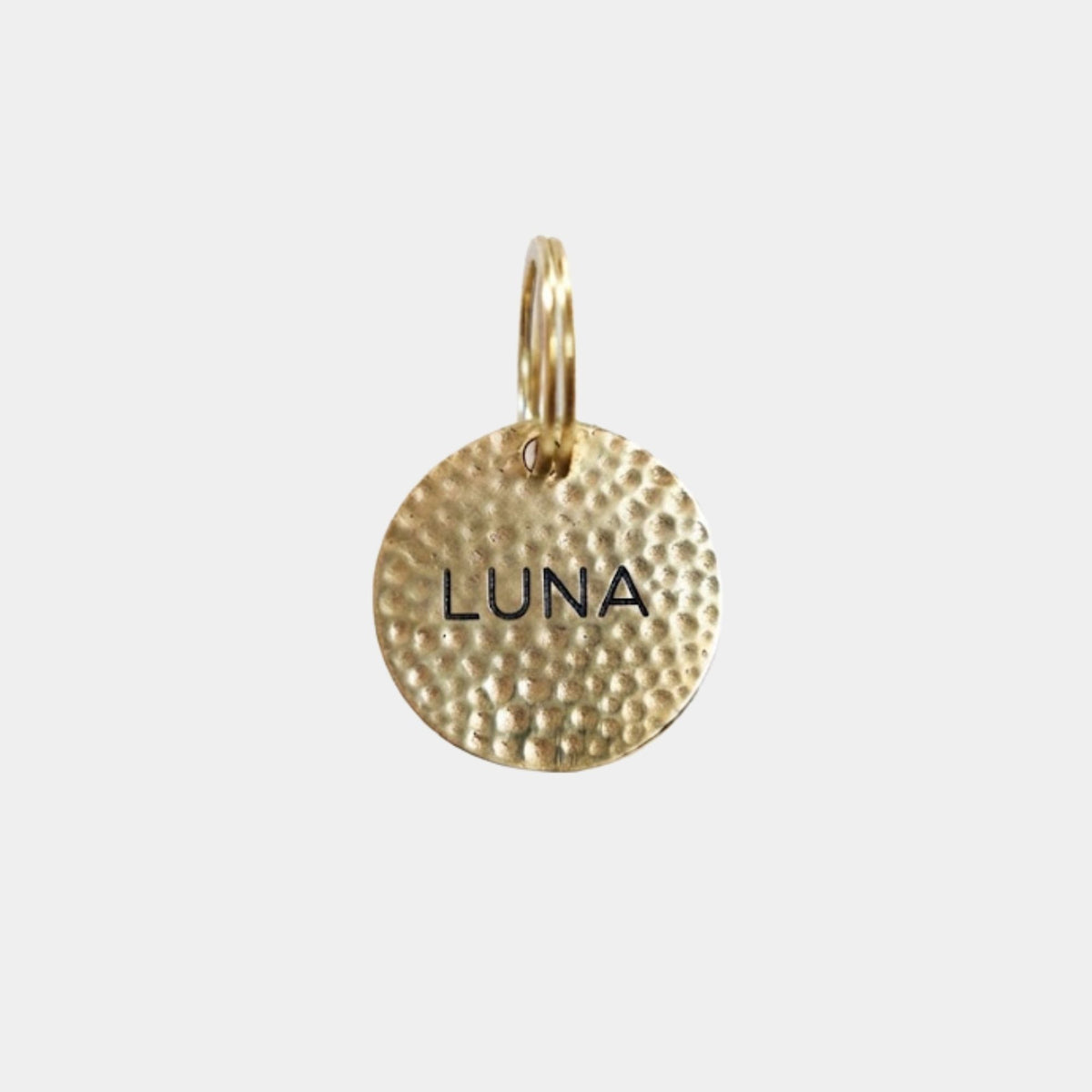 A gold-toned textured circular charm pendant with the name 'LUNA' engraved in black capital letters, shown on a light gray background.