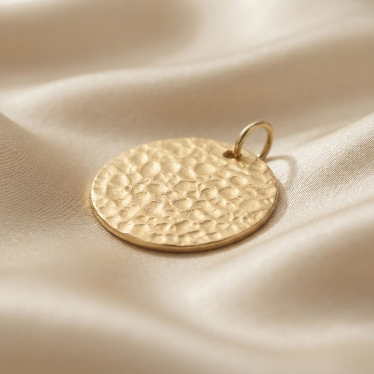 A round gold pendant with a hammered texture and a small top loop, resting on draped cream satin fabric.