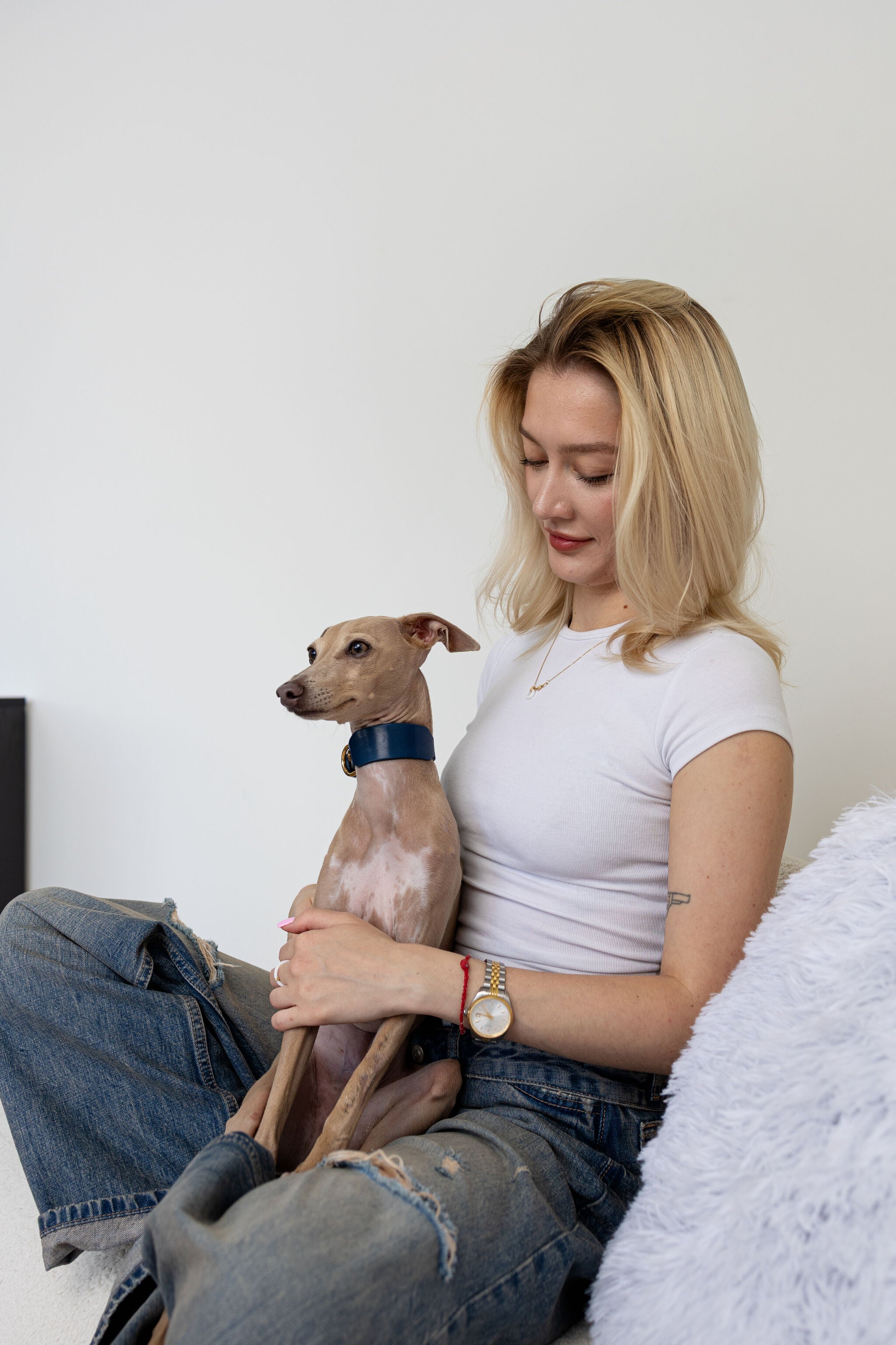 A woman with blonde hair sits on a white couch holding a small brown dog wearing a blue collar. She is wearing a white t-shirt and ripped blue jeans in a brightly lit room with a white wall.