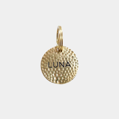 A gold-toned textured circular charm pendant with the name 'LUNA' engraved in black capital letters, shown on a light gray background.